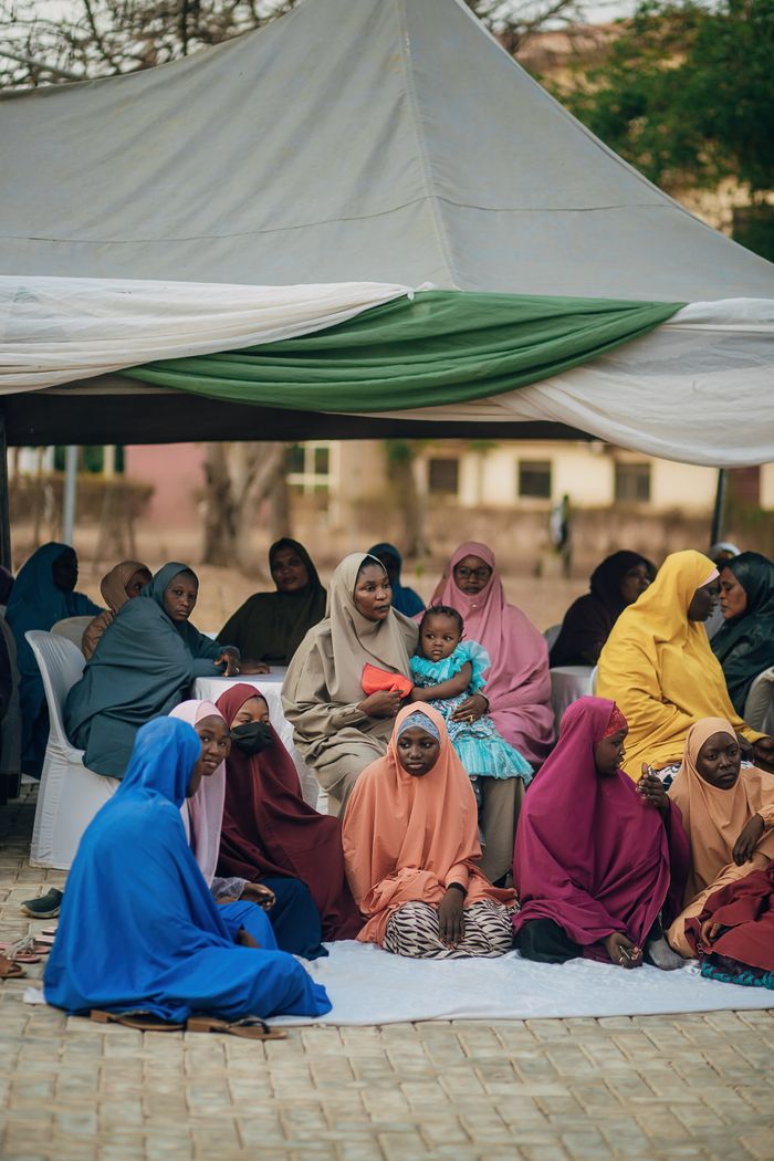 Women and children in colorful attire sit outdoors under a canopy at a community event.